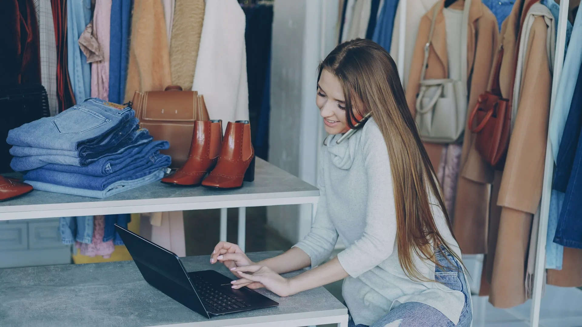 Pemilik butik bekerja di laptop di antara pakaian dan sepatu di tokonya.